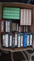 Top view of multiple 8-track tapes stacked in a cardboard box showing titles including multiple 'Sling Me a Country Song' volumes and 'Country Souvenirs'