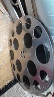 Photo shows a round aluminum film reel with multiple circular cutouts and central holes, resting on the floor at an angle, displaying the tabletop's material and design.