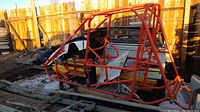 Orange tubular steel frame for a midget sprint car with some white partial skin pieces and stored outdoors on wooden pallets and ground with dirt and snow visible.