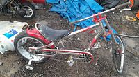 Full side view of the red and chrome Schwinn Stingray bike with training wheels on gravel and mud ground showing the overall condition.