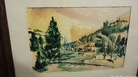 Watercolor painting showing a rural scene with trees, a farmhouse, snow fence, and hills. The painting is mounted under glass in a frame.