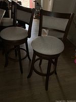 Three wooden bar stools with light beige upholstered backrests and seats, showing dark wood frames and swivel seats.