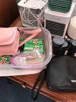 Plastic storage bin containing pink Instax Mini camera case, packs of Fujifilm Instax Mini instant film, instruction booklets, accessories in zip bag, and Tasco binoculars with black case on table.