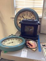 Overview of four clocks: two blue wall clocks, one black wood mantle clock, and one pink Sharp alarm clock on table.