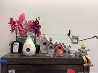 Photo of three faux orchid plants, four decorative birdhouses, and a seated bear figurine on a dark cabinet against a plain wall.