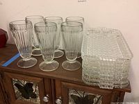 Six clear glass parfait glasses with a ribbed pattern standing next to a stack of eight clear rectangular luncheon plates with a textured geometric pattern.