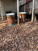 Two rusted metal paint drums outdoors amidst fallen leaves, each with a white lid, used for burning leaves and branches
