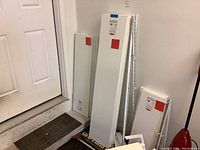 Photo showing stacked white Rubbermaid shelves in various sizes leaning against a wall near door.