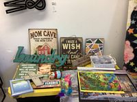Overview of multiple signs, puzzles, easels, and small photo frames on a table with various decorative wooden and metal signs including 'MOM CAVE', 'I MADE A WISH', and 'Laundry'
