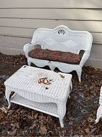 White wicker settee with patterned cushion and matching two-tiered table on an outdoor rug with autumn leaves scattered around.