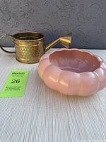 Side view of brass watering can and pink pottery planter on a light-colored textured fabric surface.