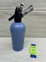 Front view of the blue metal soda syphon with black top and attached dispensing lever, sitting next to a green MaxSold lot card.