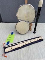 Two cream-colored recorders showing one marked AULOS with cleaning stick and cloth bag, plus two tambourines of different sizes.