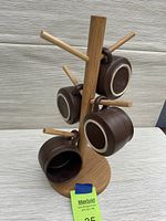 Front view of four brown ceramic mugs hanging on a wooden cup stand with a circular base and multiple horizontal pegs.