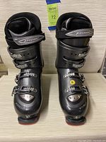 Front view of both black Atomic ski boots with four silver buckles and velcro straps on a beige fabric surface.