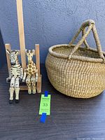 Two articulated wooden animal figurines (zebra and giraffe) sitting on a wooden frame next to a large Bolga basket on a dark surface against a textured wall.