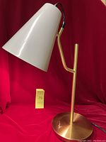 Full view of brushed gold metal desk lamp with conical off-white shade against red background. Included is a MaxSold lot tag with the number 79.