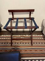 Folding wooden luggage rack with three navy blue fabric straps, standing on a patterned rug with white walls in the background.