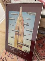 Framed vintage lithograph poster of skyscraper and zeppelins, titled 'ART EXPO NEW YORK' shown standing against wall on carpeted floor.