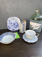 Image showing the full group of pottery and glass items on a surface against a gray wall, including the hand warmer flask, wrapped vase, teacup with saucer, bowls, and plate.