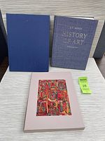 Photo of three hardcover art books displayed on a white tablecloth: 'History of Art' by R.W. Janson with textured grey cover, a blue hardcover book, and a third book in a pink slipcover with colorful abstract artwork.