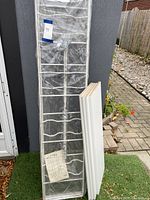 White wire shelving unit frame wrapped in plastic with attached IKEA Enetri manual, alongside 4 stacked white shelves leaning against it outdoors.