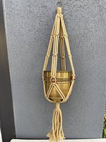 Full view of beige macrame plant hanger holding a brass planter against a gray textured wall.