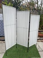 Three-panel metal folding screen with white sheer fabric panels standing outdoors on a stone patio.