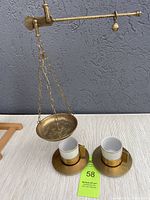 Photo showing brass Roman market scale with hanging basket alongside two brass demitasse cups with white ceramic inserts on matching brass saucers on a flat surface.