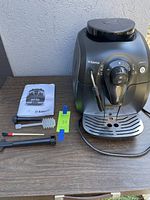 Front and side view of the black Saeco espresso machine with milk frother wand and control dial visible, alongside included accessories and user manual on the table.