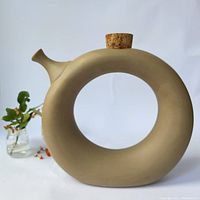 Front view of circular ceramic wine tote with cork stopper and spout, placed next to a small plant.