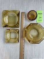 Four amber glass ashtrays of various shapes and sizes including square, octagonal, and round, with a ruler for scale.