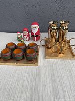 Photo showing Christmas-themed stacking (nesting) dolls, wooden plaid napkin rings on brass tray, gold trays with handles, and two gold glass vases.