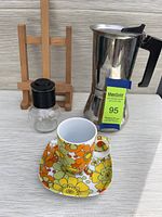 Photo showing the XL Vev stovetop espresso maker, spice jar with black lid, colorful cup and matching saucer, and a wooden stand in the background.