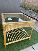 Full view of vintage rattan coffee table with glass top on outdoor artificial grass, showing square shape and open lower shelf.