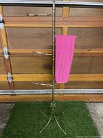 Full view of the vintage brass-effect standing towel rack with four arms and a pink towel hanging on one arm. Shows tripod base and overall height in context with garage door.