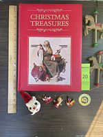 Photo of red hardcover book titled Christmas Treasures by Deborah Cannarella, showing padded and slightly worn cover. Assorted holiday ornaments arranged below including honeycomb paper Santa, miniature wooden ornaments, and a thin gold metal ornament.