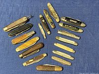 Eighteen assorted vintage folding pocket knives laid out on blue fabric showing various handle materials and shapes.