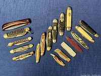 Full view photo showing 18 penblades of various sizes, handle styles, and colors laid out on blue cloth surface.