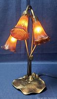 Front angle corner view of the lamp illuminated, showing three bell-shaped orange glass shades hanging from curved brass-tone arms with a lily pad shaped base.