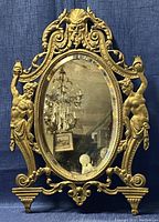 Front view of oval brass frame with beveled glass mirror, decorative cast figures and acanthus scrolls against blue background