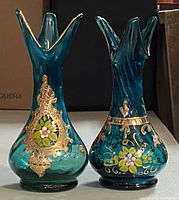 Pair of teal blue blown glass vases with flared star-shaped tops, gold painted floral and ornate decoration on the body. One vase has a framed floral medallion, the other features a bird motif with gold scrollwork.