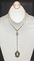 Full view of necklace and brooch shown on white bust display. Necklace is a lariat style with long chain and pendant cameo. Brooch is oval cameo with gold-tone border.