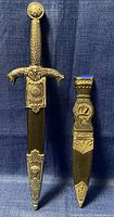 Photo of two replica daggers sheathed; King’s Arthur dagger with textured handle and lion motifs, and smaller Ireland Rampant Sgian Dubh with ornate sheath and top blue detail.