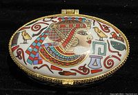 Close-up of the top side of the oval trinket box showing the hand painted Egyptian profile and motifs on a white background with a gold-tone metal trim.