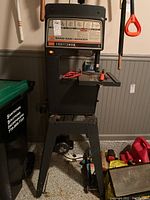 Front view of the black and grey Craftsman 12 inch Band Saw-Sander with metal stand, placed indoors near garage tools and trash bin.