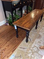 Side view of wooden bench showing black painted turned legs and natural wood seat with a small signed number tag visible.