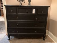 Front view of black wooden chest of drawers showing six drawers and ring pulls, standing against beige wall on concrete floor.