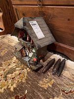 Full view of the wooden chalet Lotscher cuckoo clock with roof, weights, key, and chains visible on the carpet floor. Shows scale and included accessories.