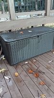 Side view of black plastic deck box on wooden deck with leaves around, showing woven pattern